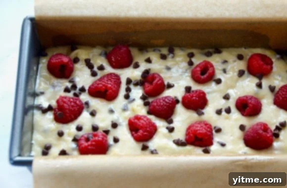 A loaf pan filled with banana bread batter topped with raspberries and chocolate chips before baking, illustrating the layering technique.