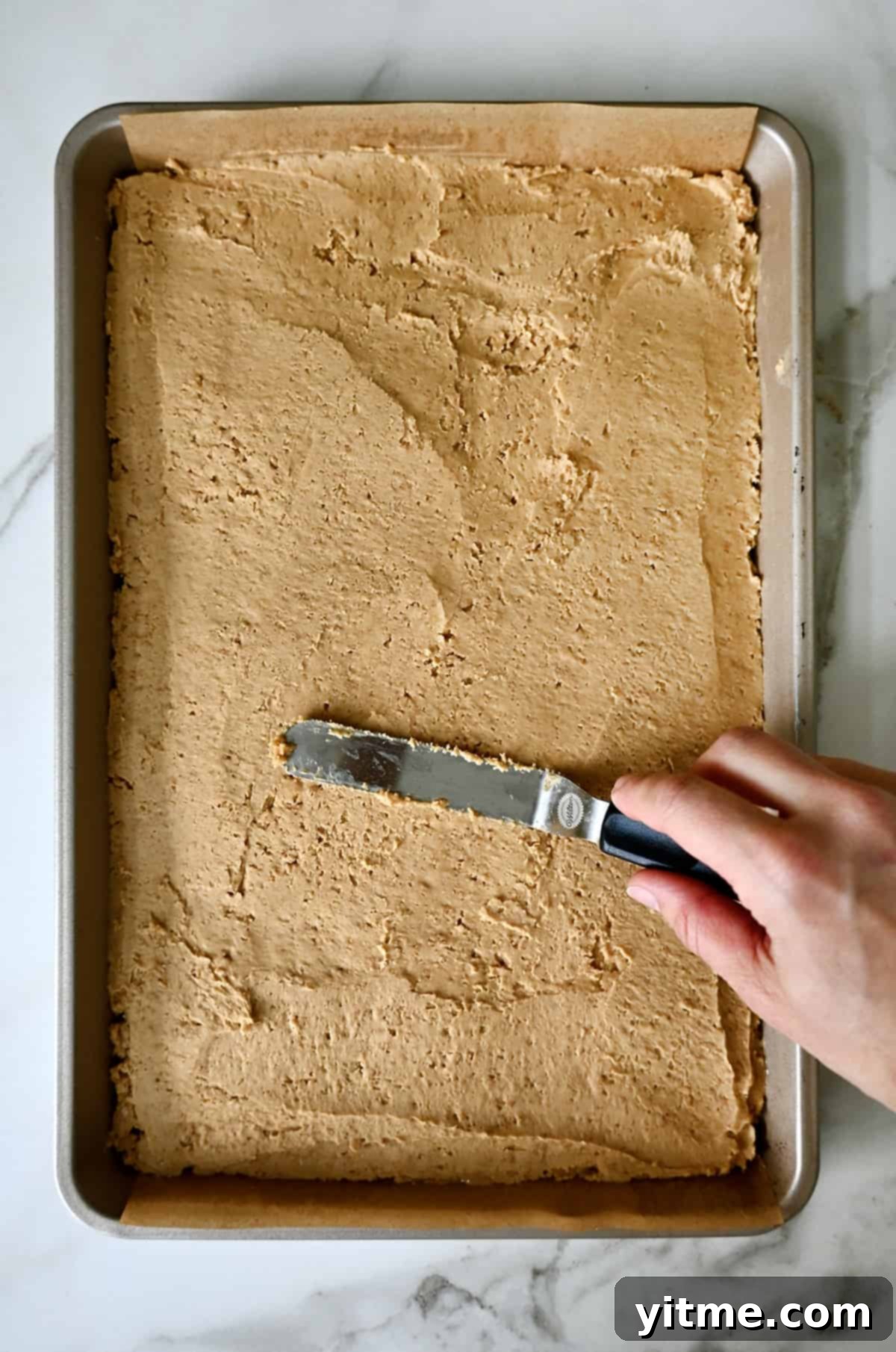 Spreading peanut butter bars evenly in a baking pan with an offset spatula.