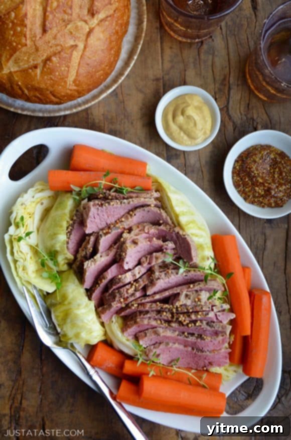 White plate containing The Best Slow Cooker Corned Beef and Cabbage with carrots next to a loaf of bread and two small bowls containing mustards