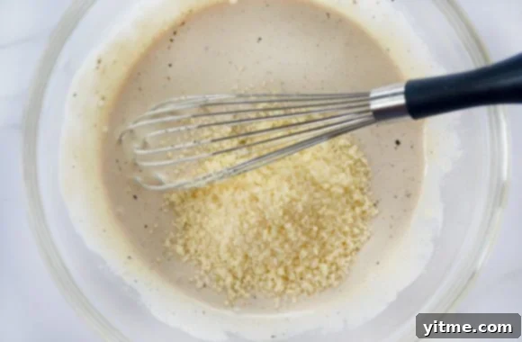 Glass bowl containing caesar salad dressing, parmesan cheese and a whisk