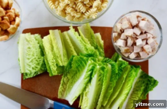 Chopped romaine on cutting board with knife next to three bowls containing chicken, pasta and croutons