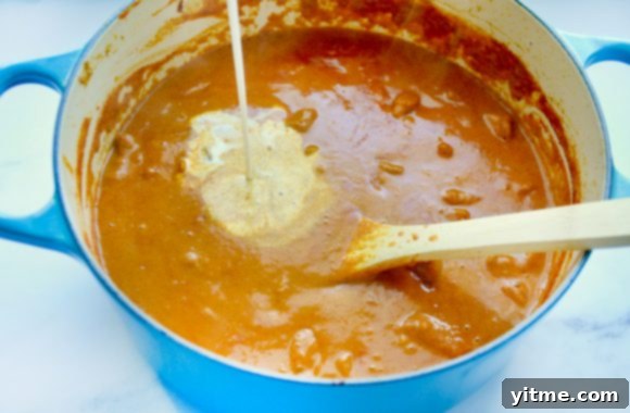 Irresistible Chicken Tikka Masala 4 A stream of rich heavy cream being poured into a blue enamel saucepot, mixing into a simmering, deeply colored curry sauce. A wooden spoon rests inside the pot, suggesting active preparation of the Chicken Tikka Masala sauce.