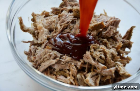 Fork-Tender Instant Pot BBQ Pulled Pork 4 Clear bowl containing pulled pork with barbecue sauce being added