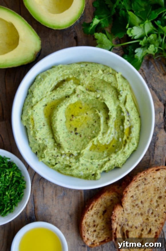 A white bowl containing avocado hummus with toast and herbs around it