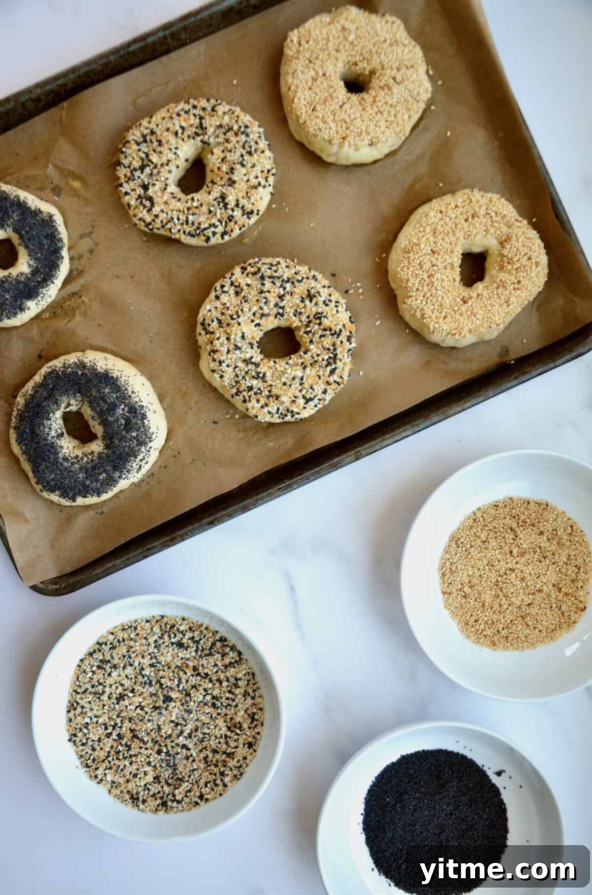 Unbaked homemade bagels on a baking sheet with three different toppings: poppyseed, sesame and everything bagel.
