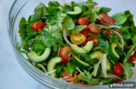A glass bowl filled with fresh Thai salad ingredients: crisp cucumber, sliced shallots, fragrant mint, vibrant cilantro, and juicy cherry tomatoes.