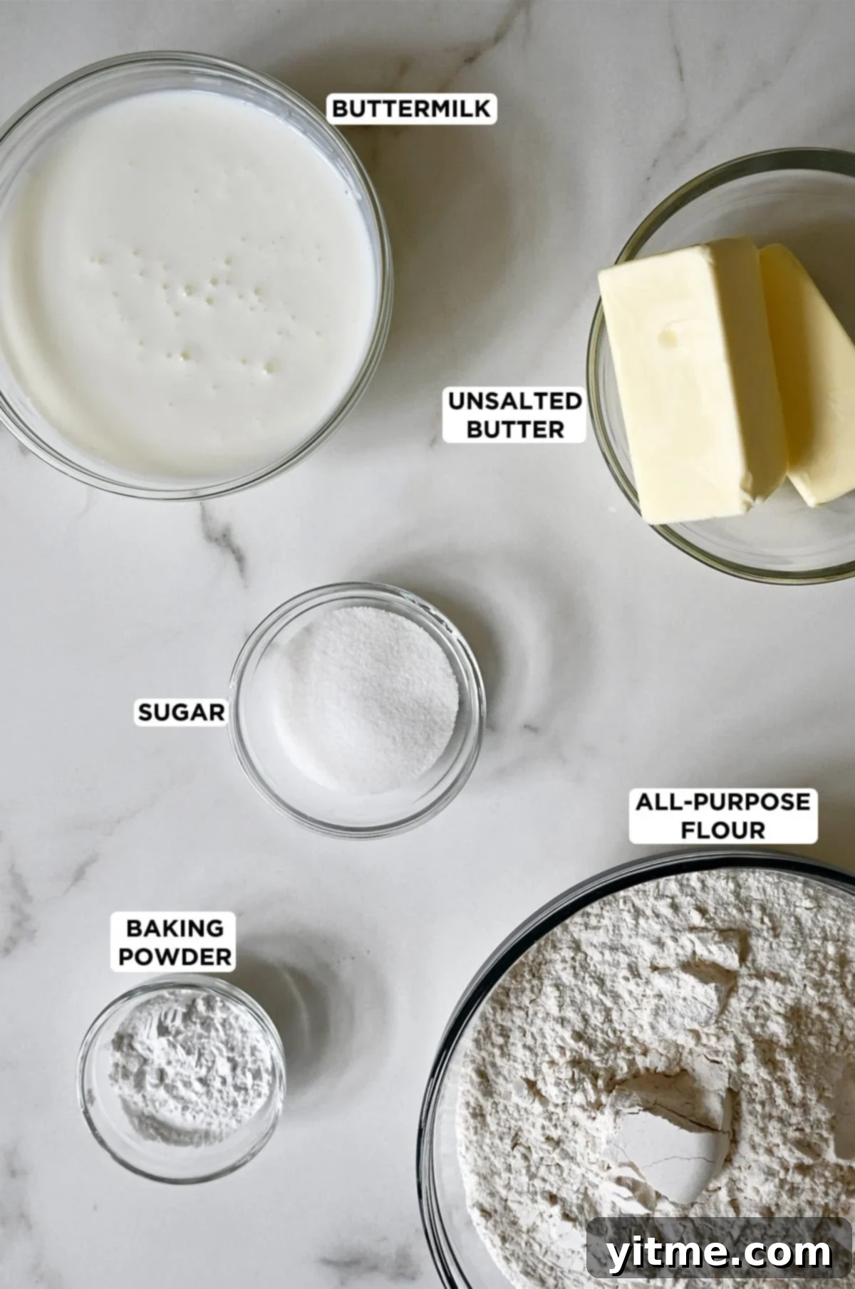 Buttermilk biscuit ingredients in glass bowls, including flour, baking powder, sugar, butter, buttermilk and salt.