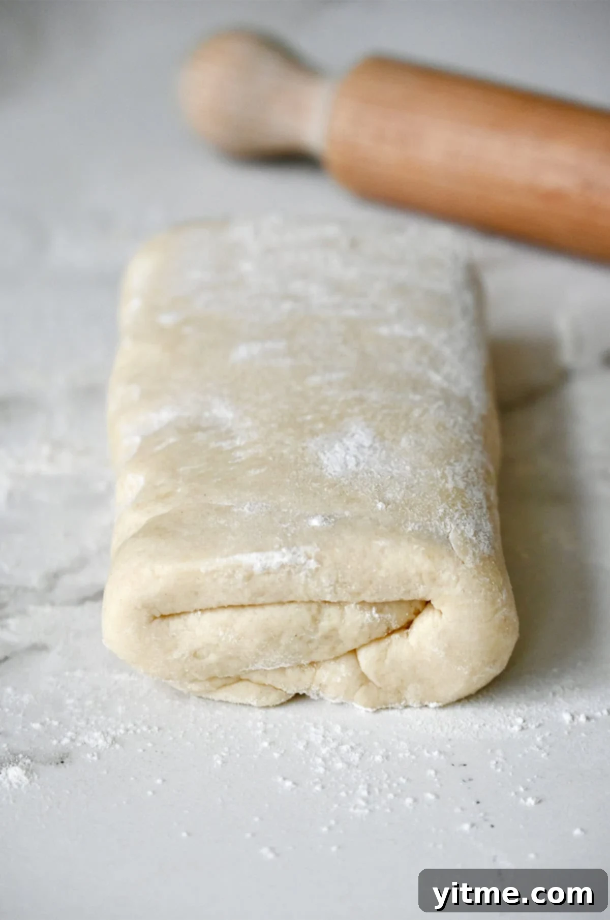 Folded biscuit dough on a lightly floured surface next to a rolling pin.