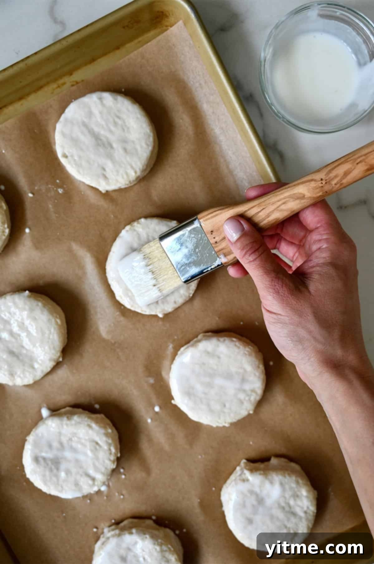 Brushing buttermilk atop biscuit dough rounds on a parchment paper-lined baking sheet.