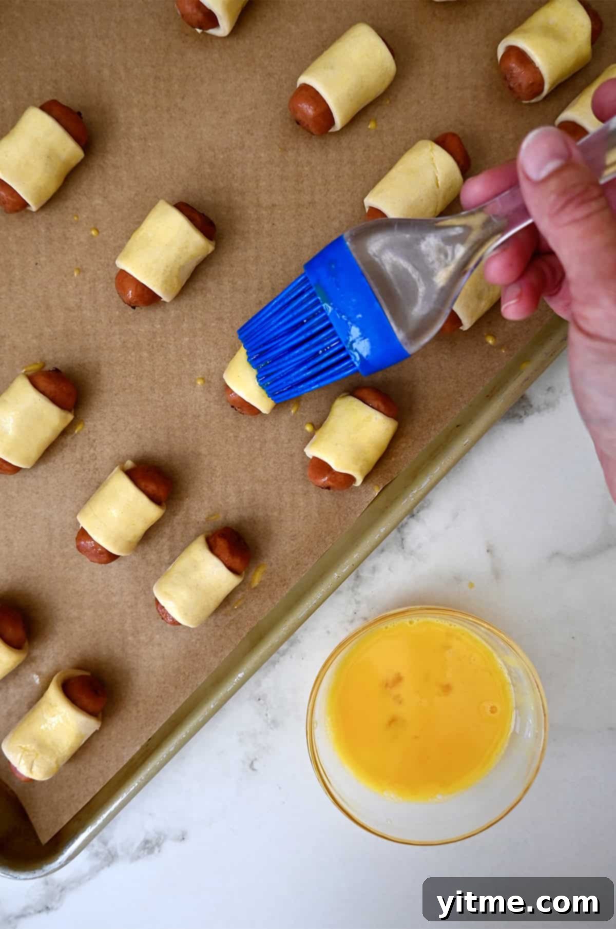 Using a pastry brush to add an egg wash on top of unbaked pigs in a blanket on a baking sheet.