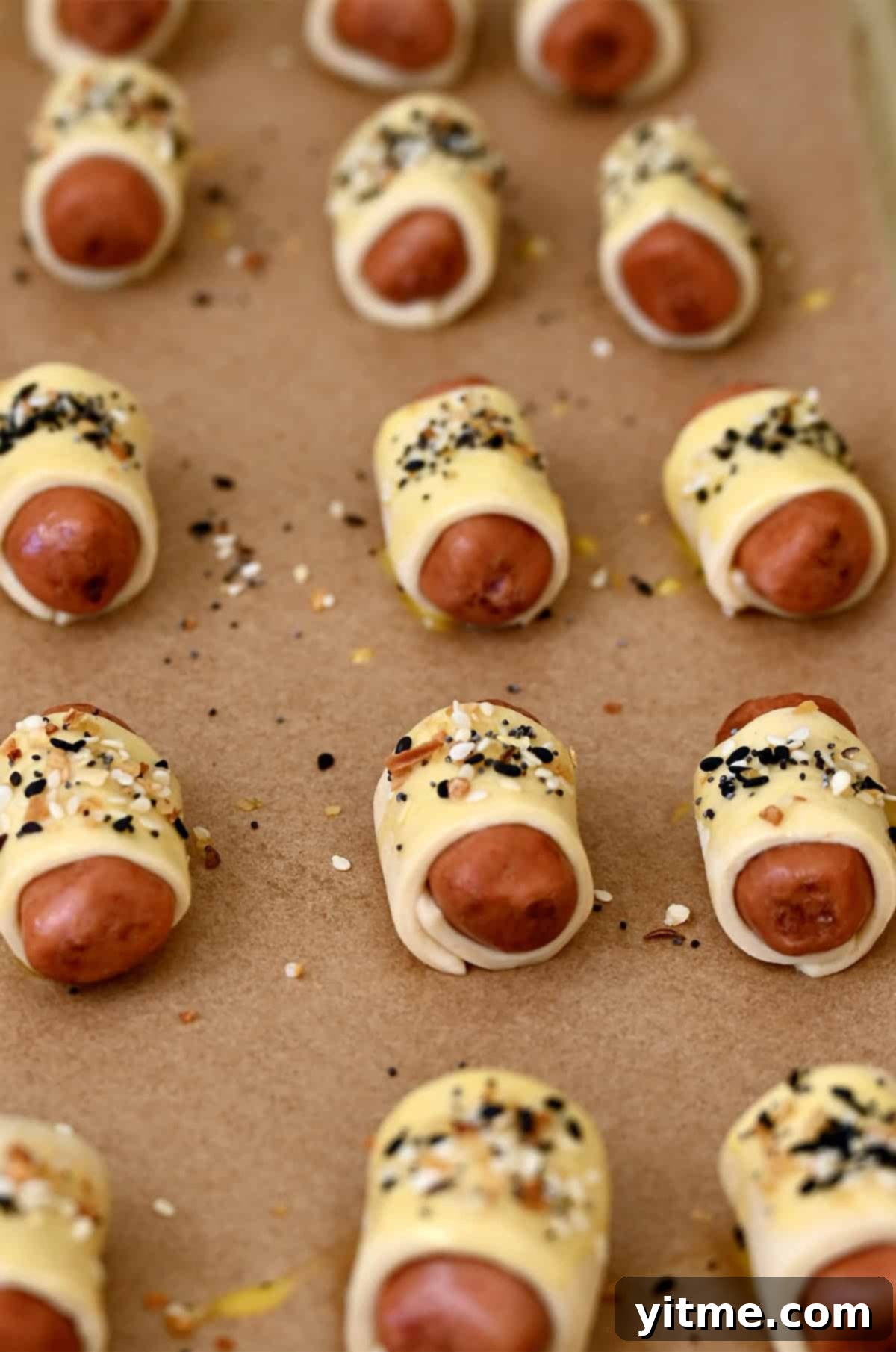 Unbaked mini pork sausages wrapped in dough and sprinkled with Everything Bagel seasoning on a baking sheet.
