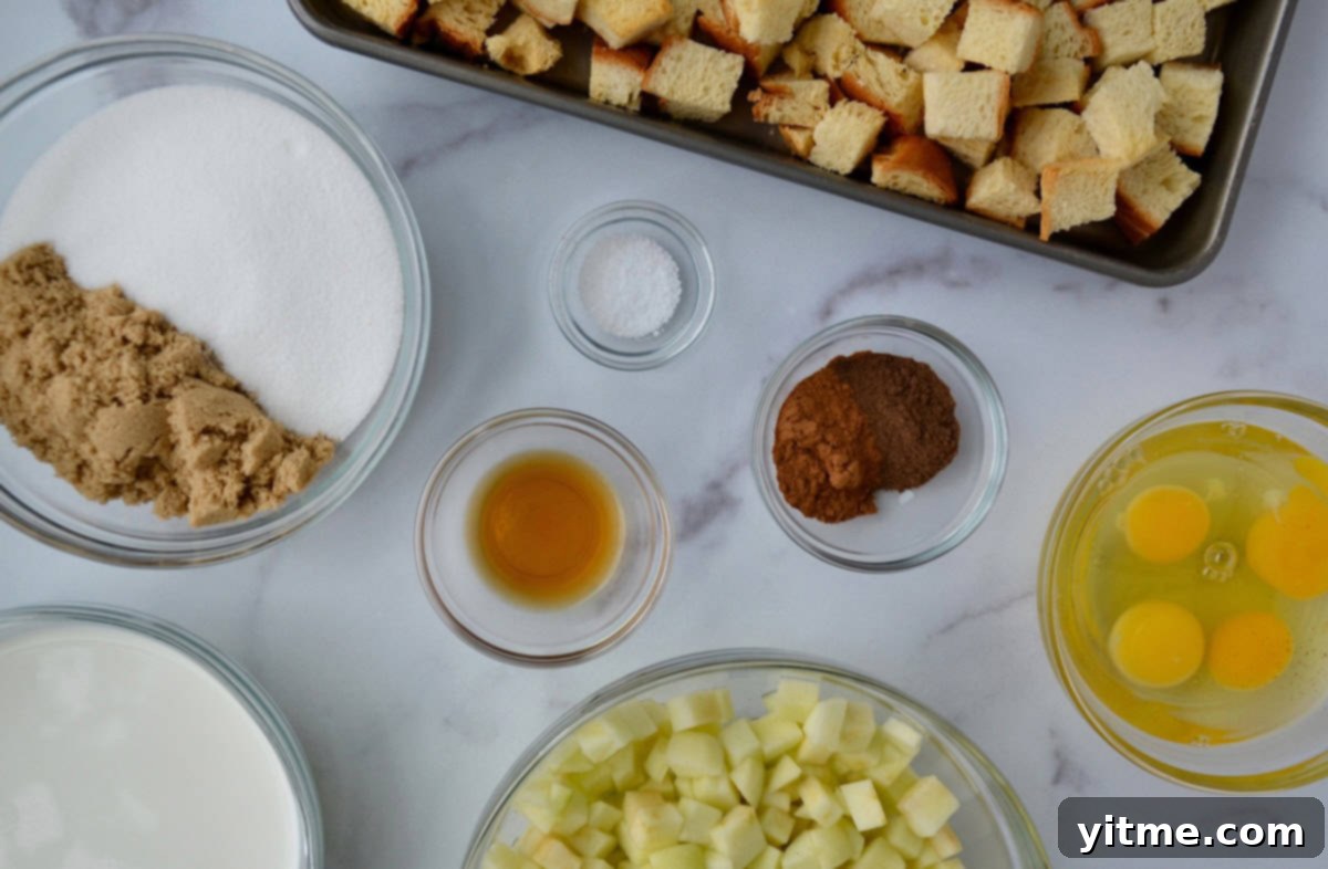 Ingredients for making apple bread pudding, including a baking sheet with cubed brioche bread, next to glass bowls containing chopped apples, pumpkin pie spice, white and brown sugar, lemon juice, half-and-half, and eggs.