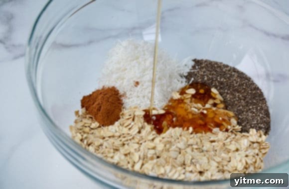 A glass bowl containing oats, coconut, spices and honey ready to be mixed