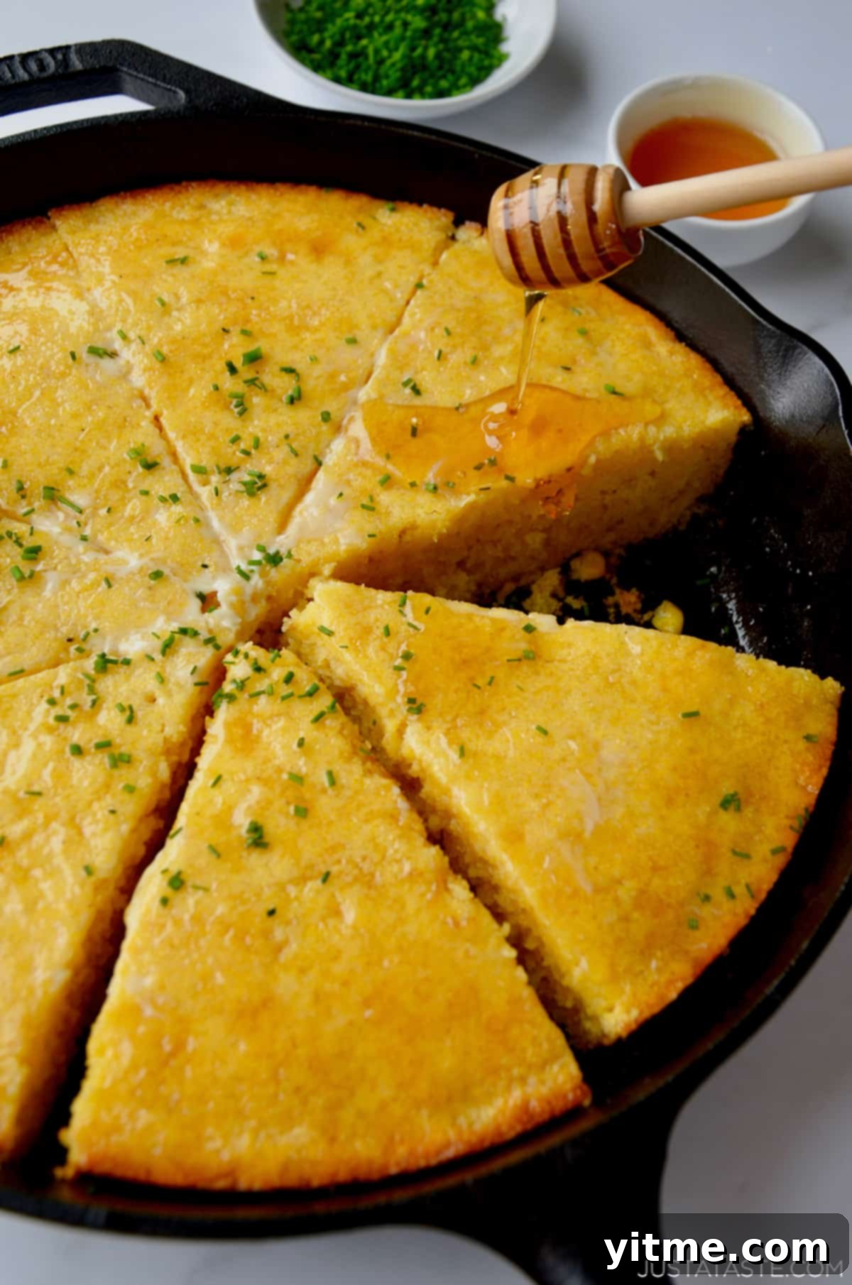 Honey being drizzled over freshly sliced cornbread in a cast iron pan. The cornbread is beautifully golden, topped with a dollop of melting butter and a sprinkle of minced chives. Small decorative bowls containing more chives and honey are arranged beside the rustic skillet.