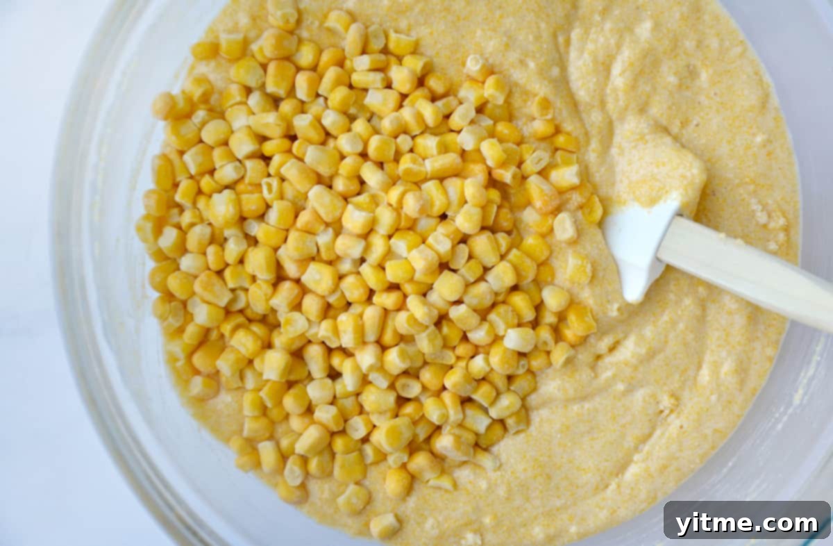 A glass mixing bowl filled with creamy cornbread batter. Fresh or frozen corn kernels are visible on top of the smooth batter, and a rubber spatula rests against the side of the bowl, ready to finish mixing.