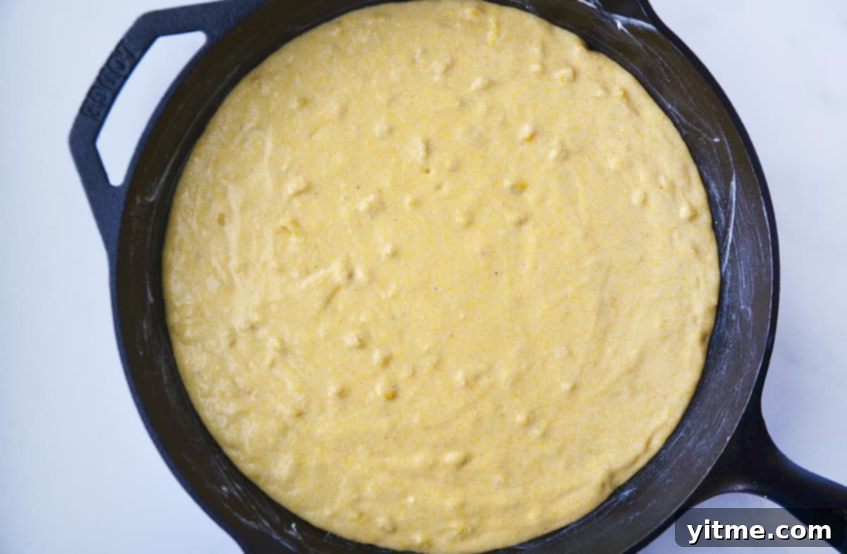 Smooth, yellow cornbread batter has just been poured into a seasoned cast iron skillet, ready for baking. The batter is evenly spread, indicating a perfect preparation for a golden crust.