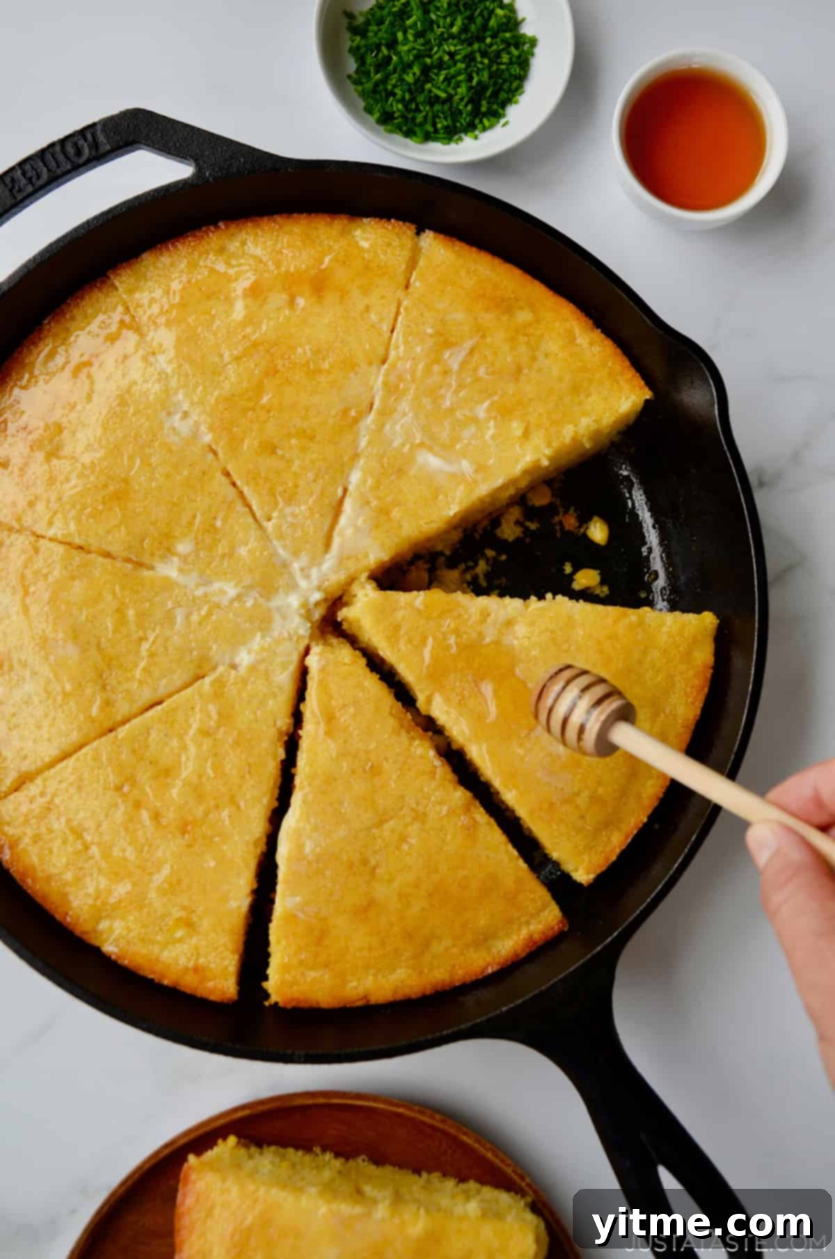 A person is gracefully drizzling golden honey over a freshly sliced cornbread, still warm in its cast iron pan. Beside the pan, a single slice of cornbread sits on a plate, alongside a small bowl of minced chives and another small bowl of honey, ready for serving.
