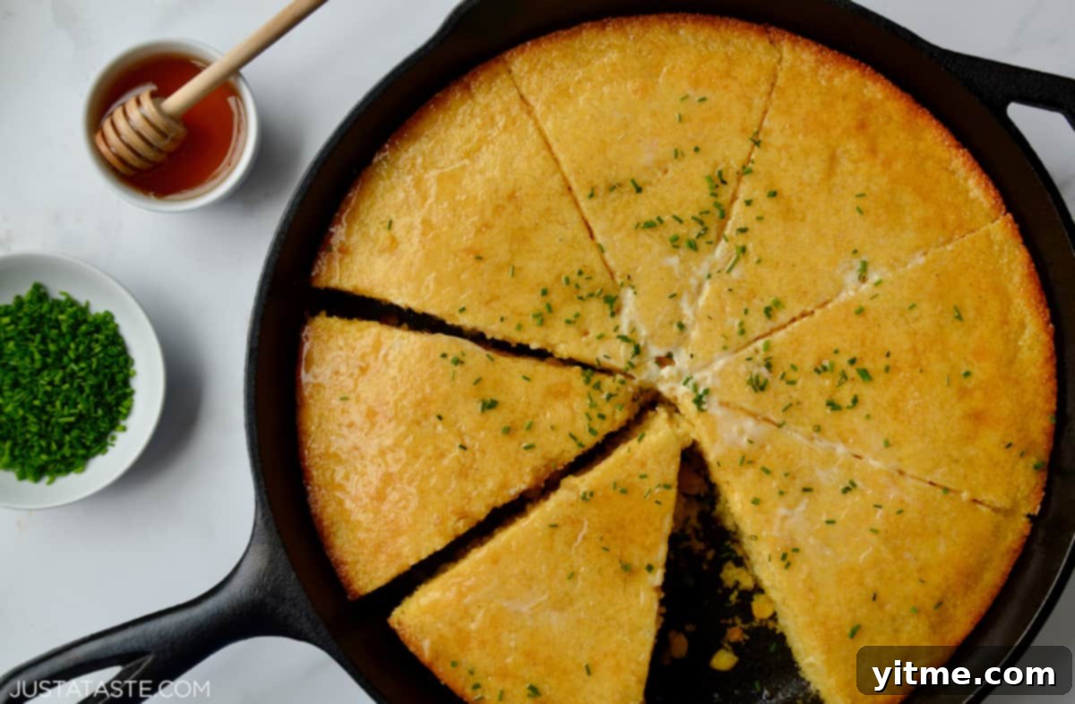 A perfectly baked and sliced cornbread rests invitingly in a cast iron skillet. It's garnished with a melting pat of butter, a scattering of vibrant minced chives, and a glistening drizzle of honey. Adjacent to the skillet are small bowls filled with additional minced chives and honey for personalized serving.