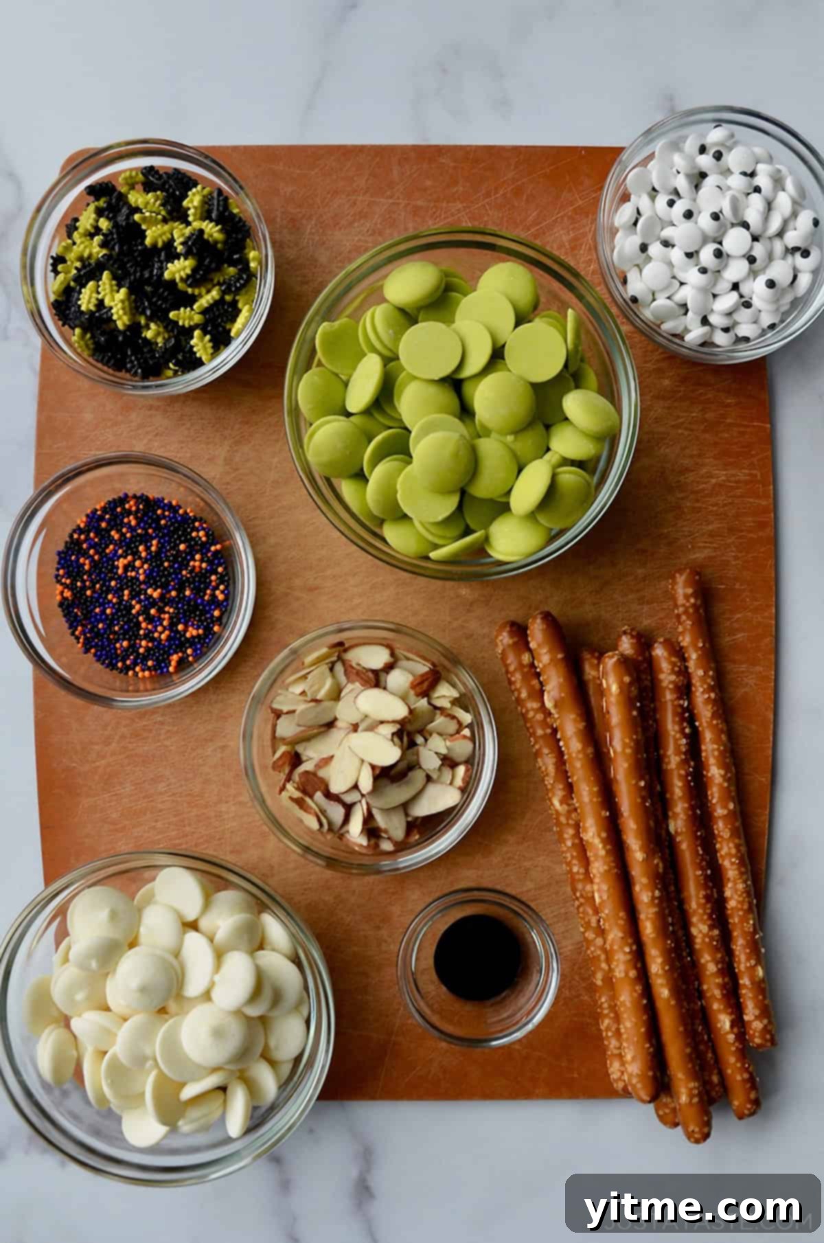 A cutting board containing glass bowls with ingredients for making chocolate-covered Halloween pretzels.