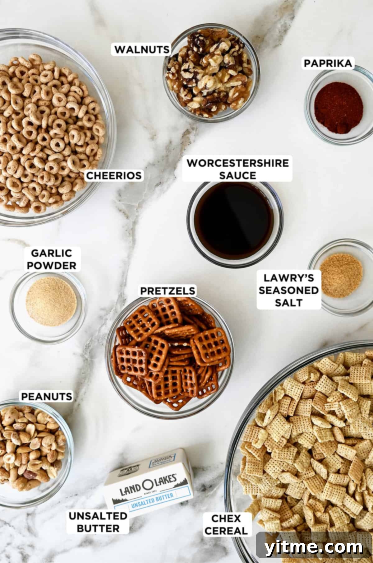 A stick of butter next to various sizes of clear bowls containing Cheerios, walnuts, paprika, Lawry’s Seasoned Salt, Chex cereal, pretzels, peanuts and garlic powder.