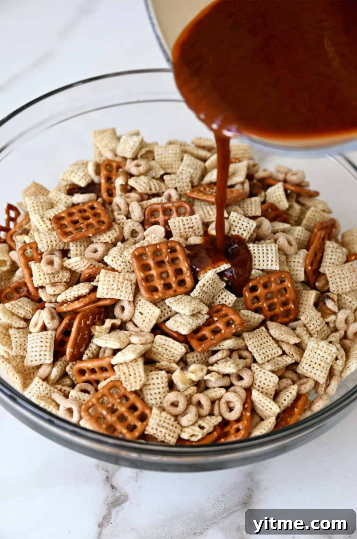 A seasoned butter mixture is being poured from a small saucepan atop Chex mix in a clear bowl.
