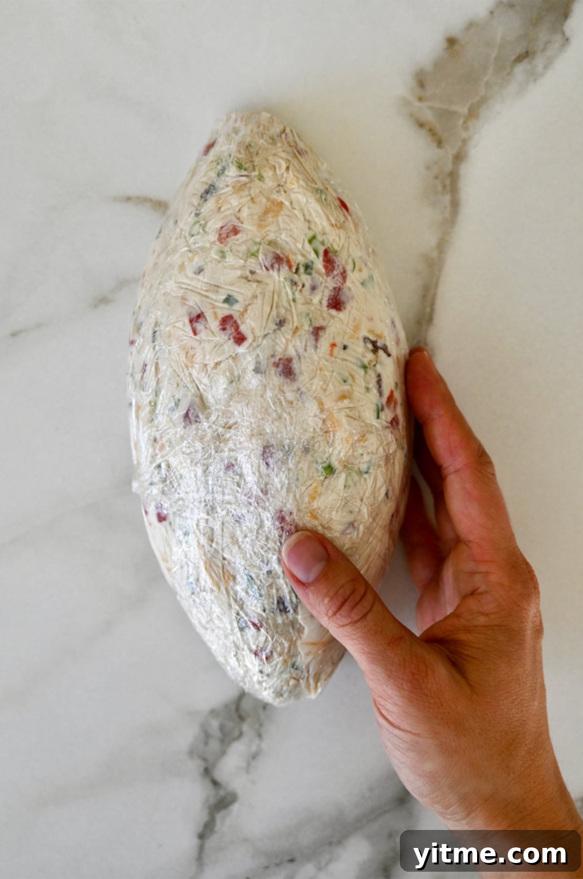 A hand holding a cheese ball shaped like a football wrapped in plastic wrap.