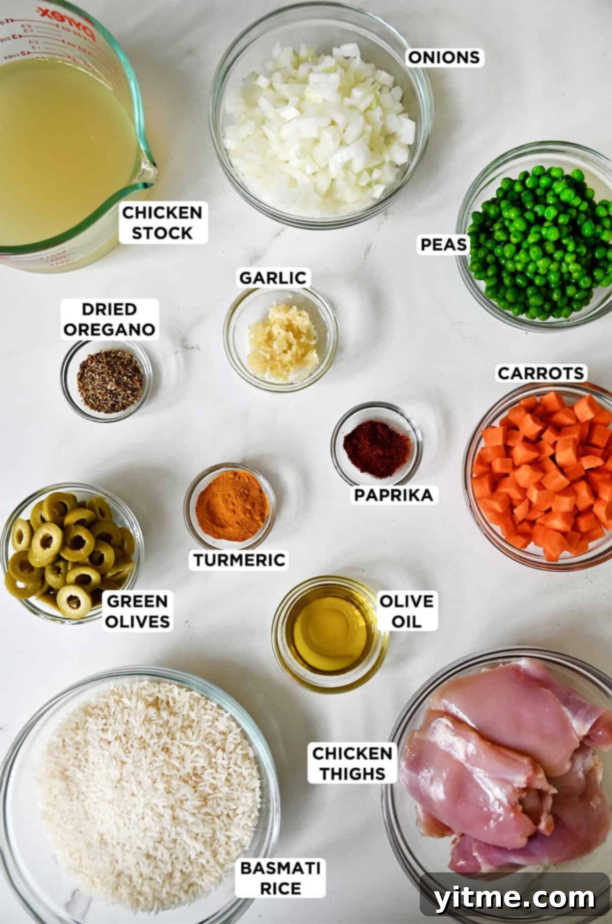 Chicken yellow rice ingredients in various sizes of glass bowls, including chicken stock, long-grain rice, peas, diced carrots and onions, paprika, olive oil, chicken thighs, turmeric, minced garlic, dried oregano and green olives.