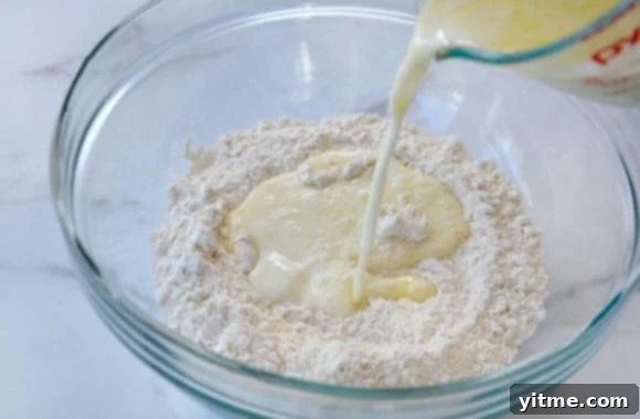 A large glass bowl containing flour with buttermilk and melted butter being poured in