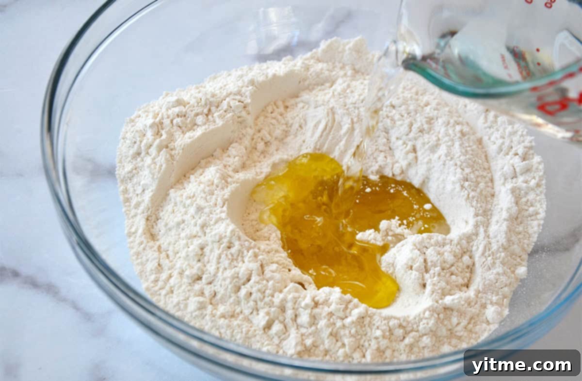 A glass bowl with all-purpose flour, and olive oil and water being poured in to begin the dough mixing process.