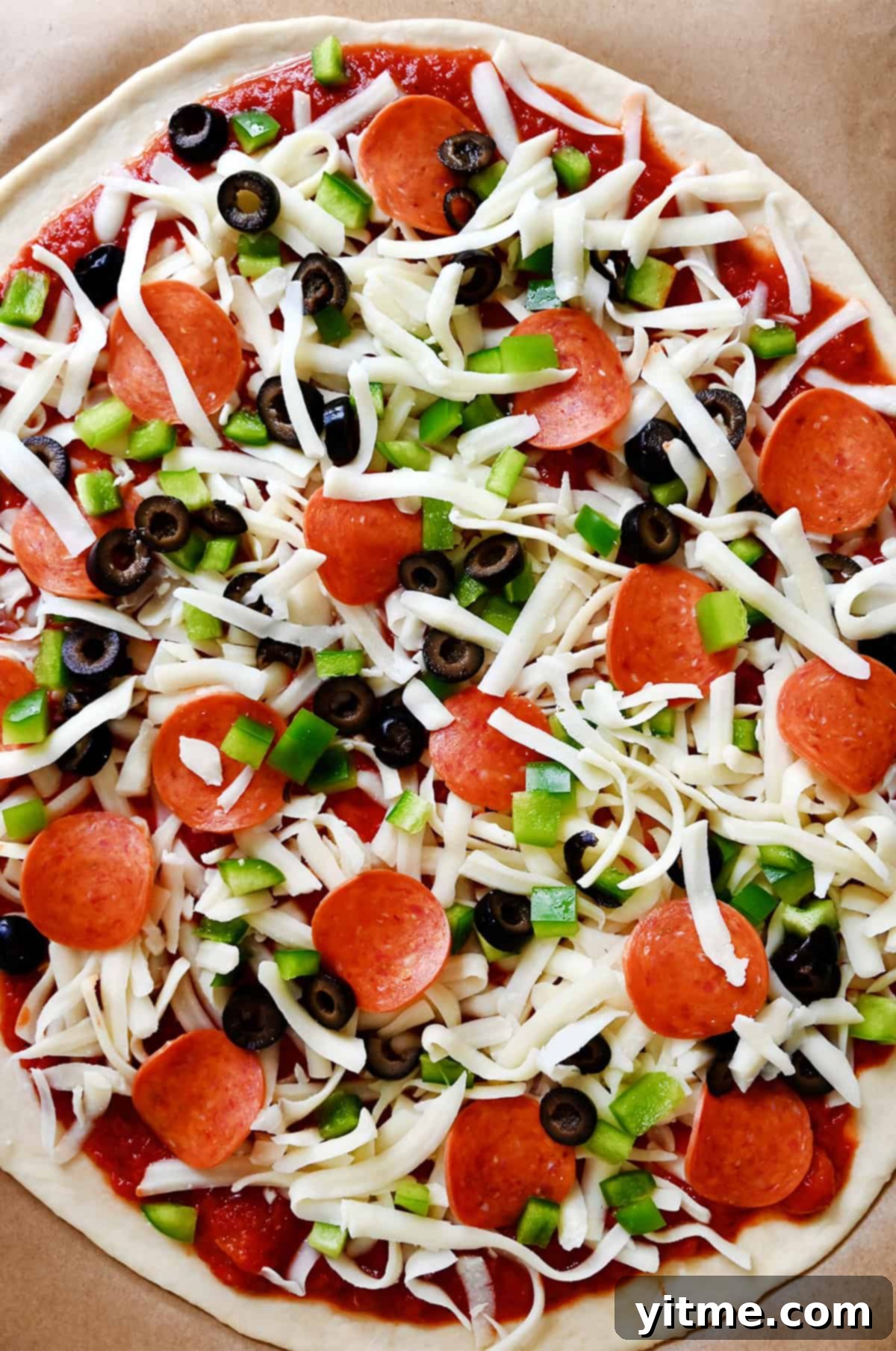 Unbaked pizza richly topped with shredded mozzarella cheese, mini pepperoni, diced green pepper, and sliced black olives, ready to enter the oven.