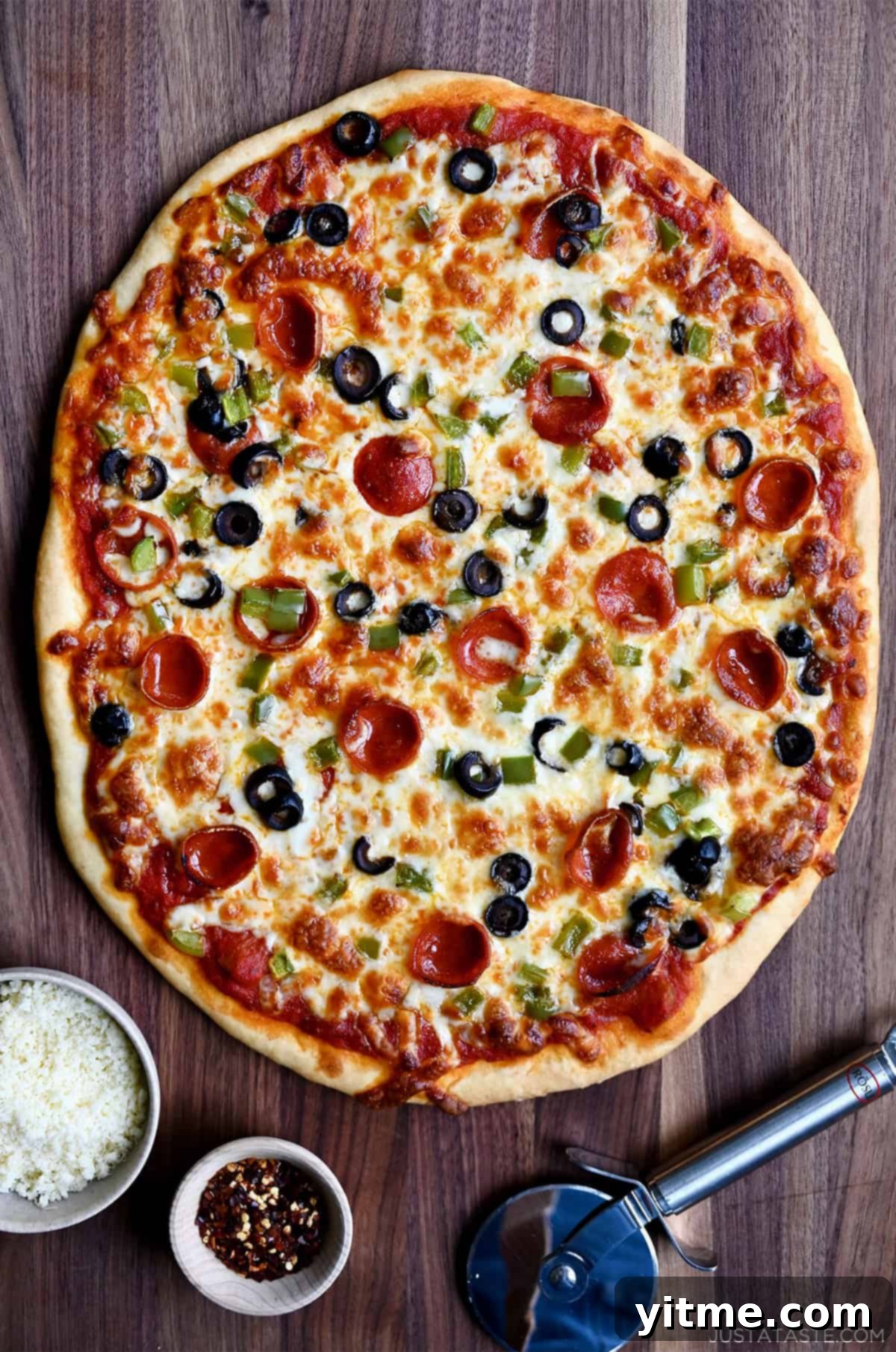 A beautifully arranged scene featuring a homemade pizza with a no-yeast crust, pizza sauce, melted cheese, mini pepperoni, diced green pepper, and black olives. Alongside the pizza are small bowls filled with grated Parmesan cheese and crushed red pepper flakes, accompanied by a pizza cutter, all inviting a delicious meal.