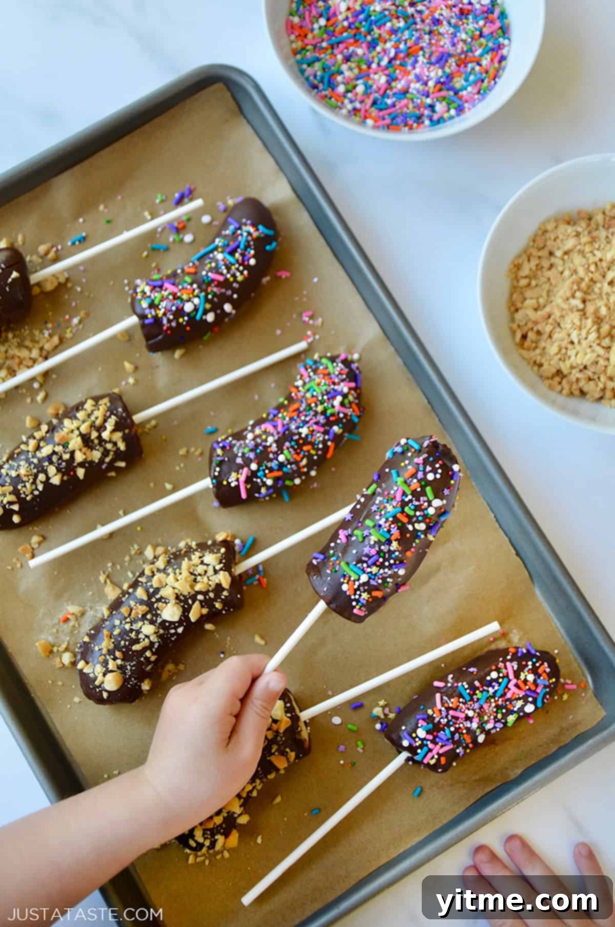 Frozen chocolate covered bananas with chopped nuts and sprinkles on a parchment paper-lined baking sheet.