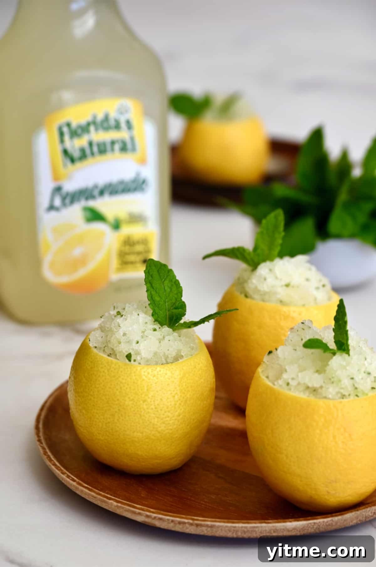 Three hollowed out lemons filled with lemon granita on a wood plate in front of a carton of Florida's Natural Lemonade.