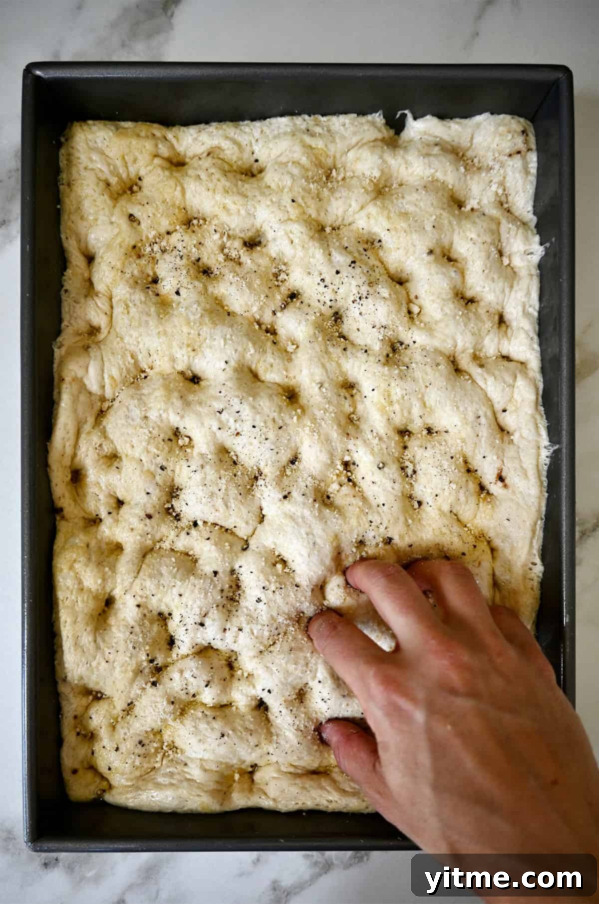 Fingers being pressed into dough in a metal baking pan, creating dimples across the surface. 