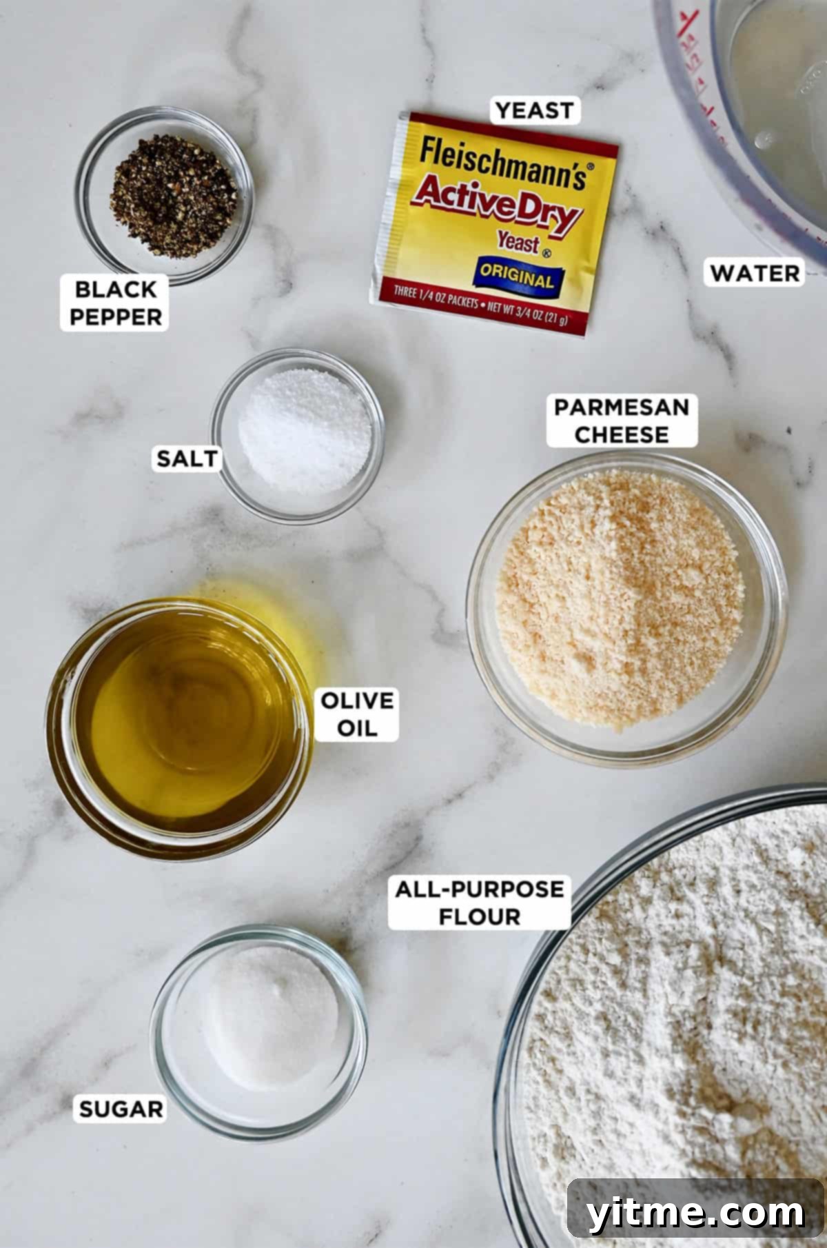 A packet of active dry yeast next to various sizes of glass bowls containing water, Parmesan cheese, flour, sugar, olive oil, salt and black pepper. 