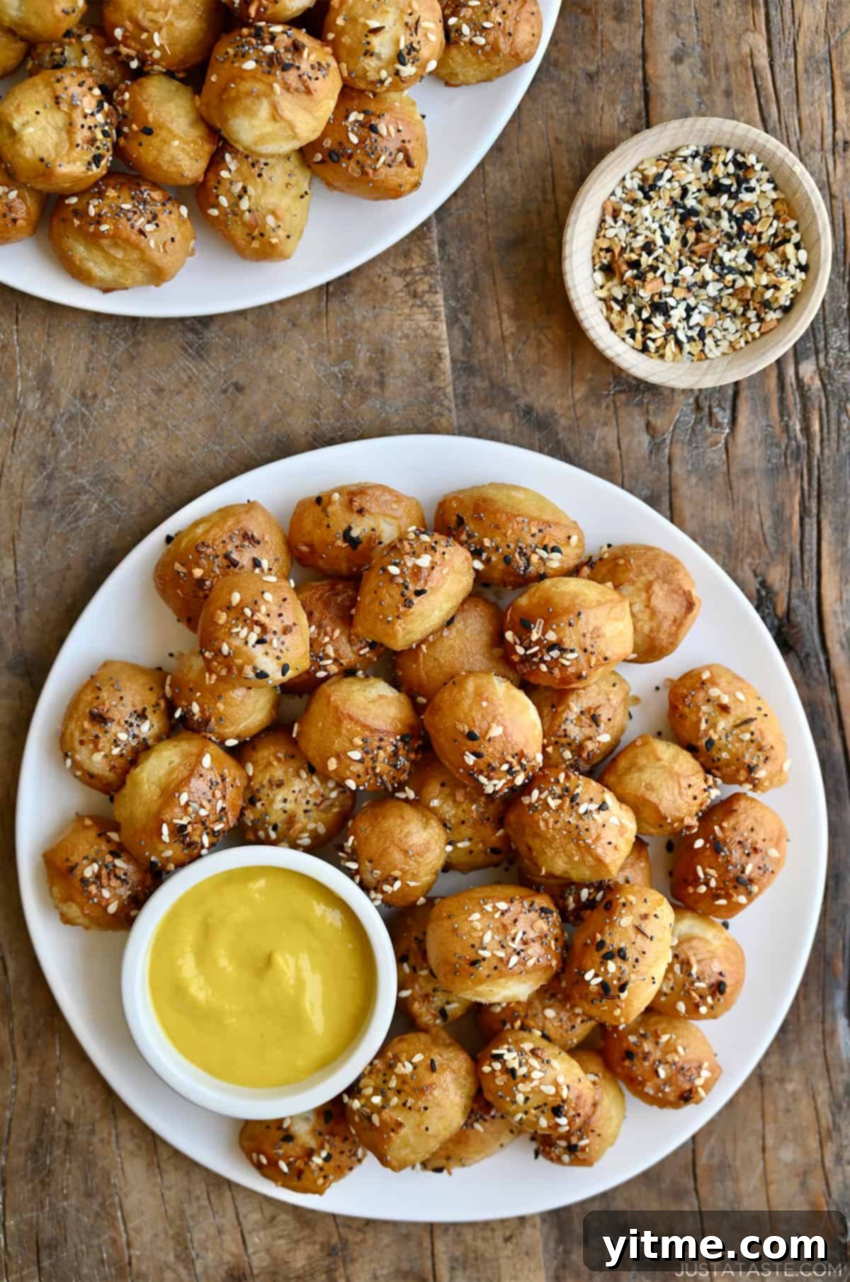 Freshly baked soft pretzel bites, seasoned with everything bagel seasoning, served on a pristine white plate alongside a small bowl of tangy yellow mustard for dipping.