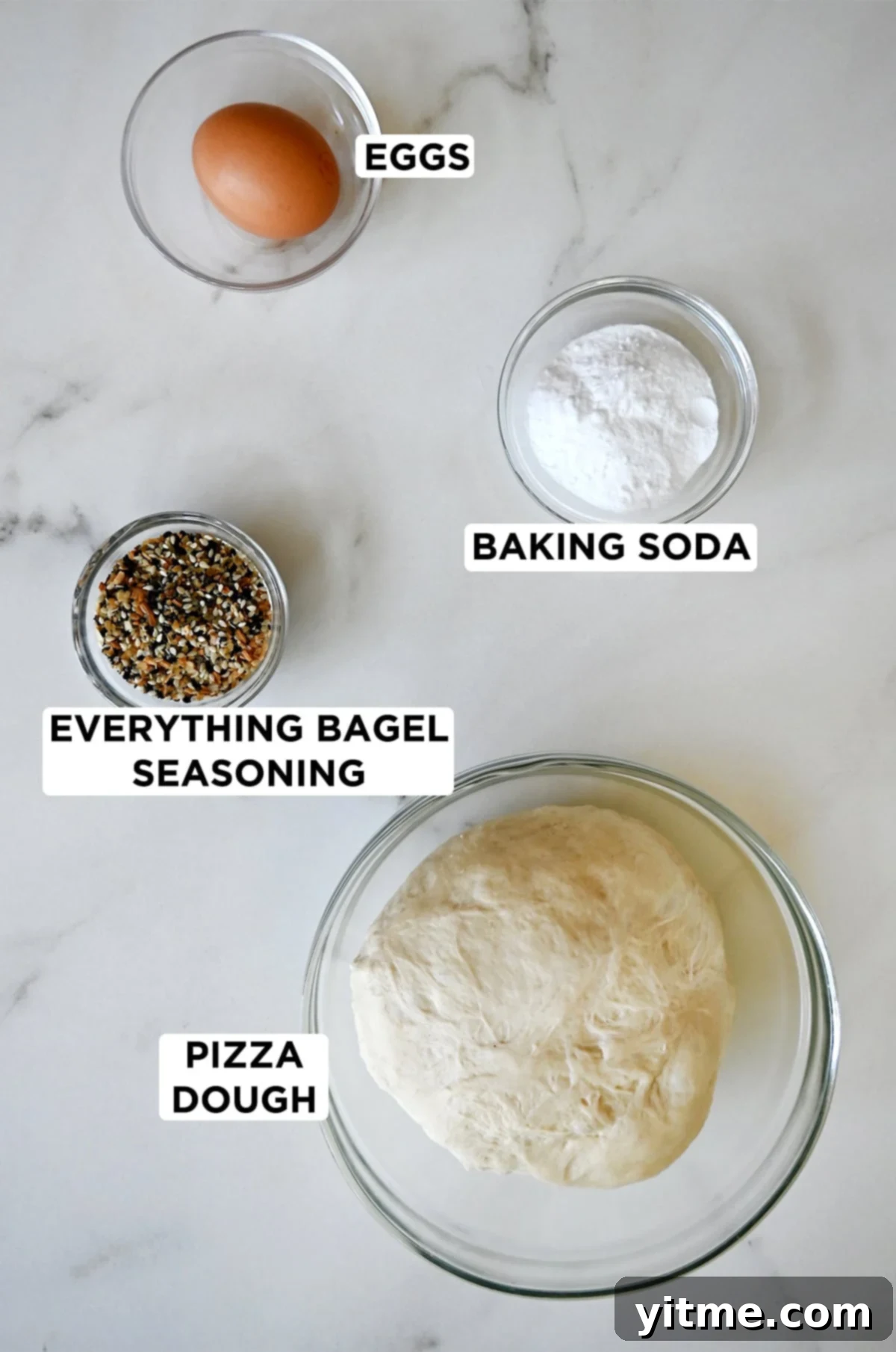 Clear glass bowls hold the essential ingredients: a single egg yolk, white baking soda, a ball of versatile pizza dough, and savory everything bagel seasoning.