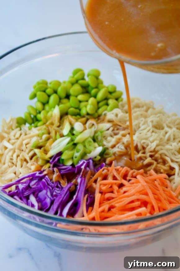 Peanut dressing being generously poured over a vibrant mix of ramen noodles, shredded carrots, purple cabbage, scallions, and edamame in a large mixing bowl.