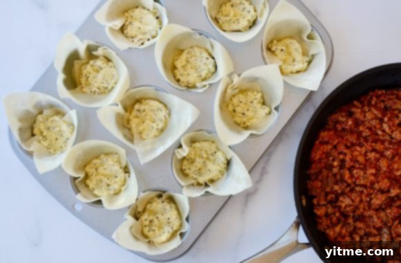Muffin pan with wonton wrappers forming cups, filled with creamy ricotta mixture, alongside a skillet of cooked Italian sausage