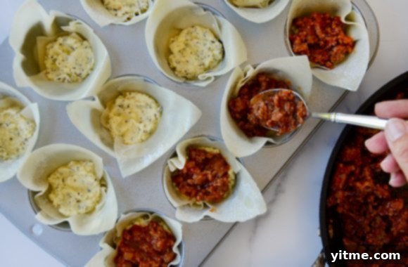 Hand holding a spoon with savory Italian sausage mixture, ready to layer into wonton cups filled with ricotta cheese