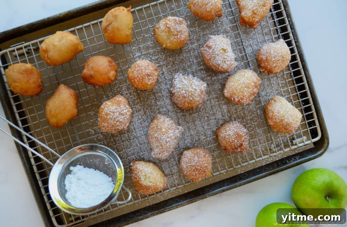 Whip Up Apple Fritters in 30 Minutes 4 Apple fritters dusted with powdered sugar on a baking sheet with a wire cooling rack.