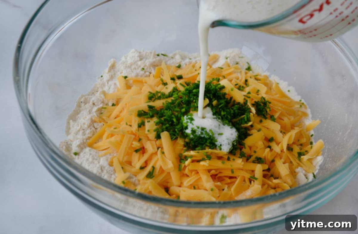 Irresistible Cheddar Biscuits 5 Buttermilk is being poured into a glass bowl containing a mixture of flour, grated butter, cheddar cheese, and chives, to form the biscuit dough.
