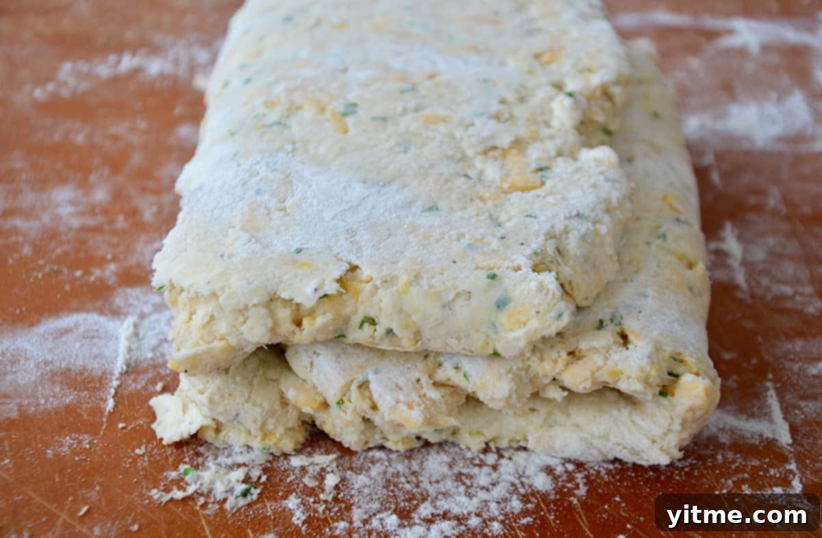 Irresistible Cheddar Biscuits 6 Layers of folded biscuit dough sit on a lightly floured wooden board, showing the preparation stage for flaky biscuits.