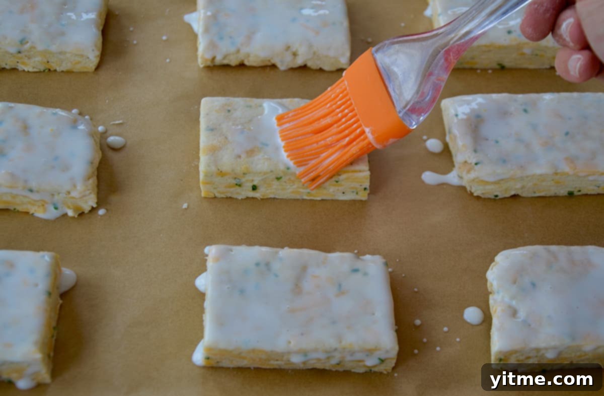 Irresistible Cheddar Biscuits 7 Rectangular unbaked biscuits on a sheet of parchment paper are being brushed with heavy cream using a silicone brush, preparing them for baking.