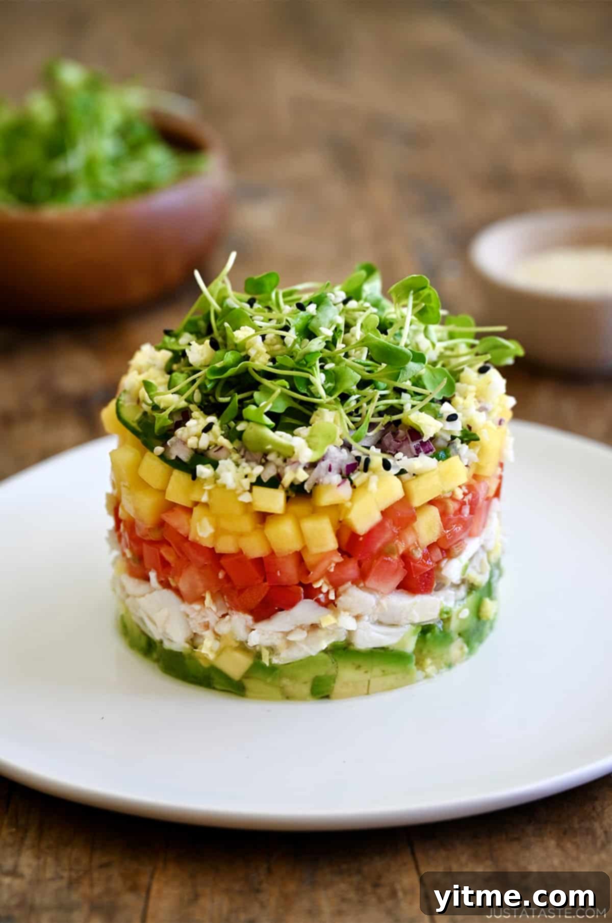 A beautifully layered La Jolla Crab Stack featuring fresh avocado, lump crab meat, ripe tomato, sweet mango, crisp red onion, and delicate microgreens, ready to be drizzled with dressing.