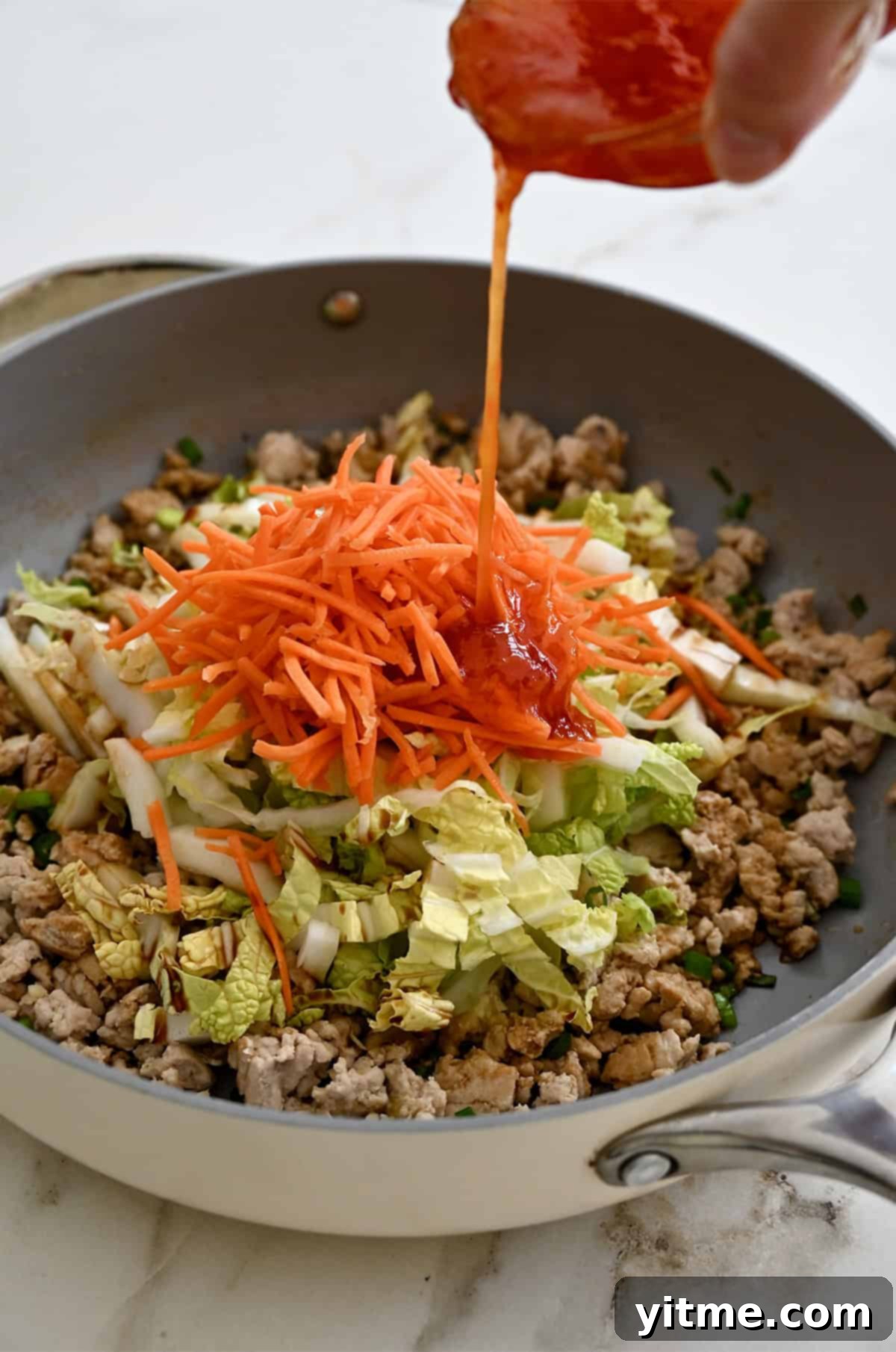 Shredded carrots, shredded cabbage and ground meat in a skillet being topped with sweet chili sauce.