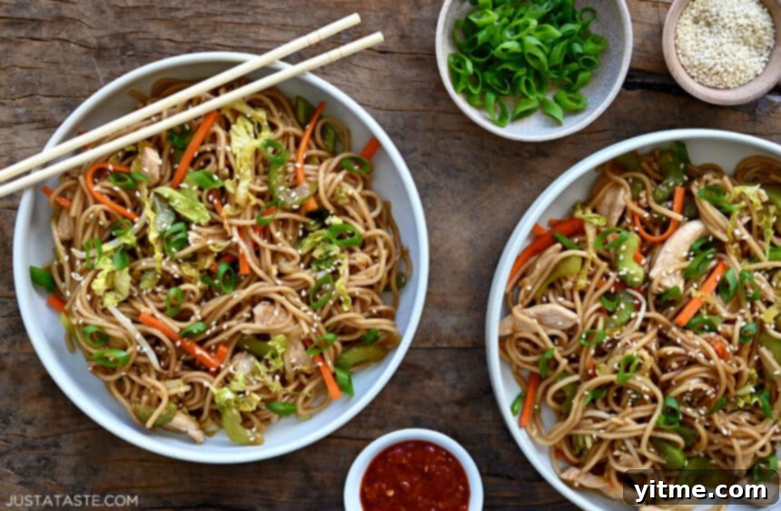 A top-down view of two bowls containing easy chicken chow mein next to small bowls containing sliced scallions, sesame seeds and chili sauce
