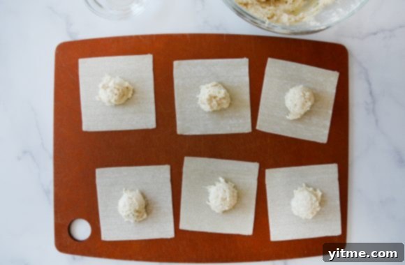 Wonton wrappers on a wooden cutting board filled with a creamy crab and cream cheese mixture, ready for folding