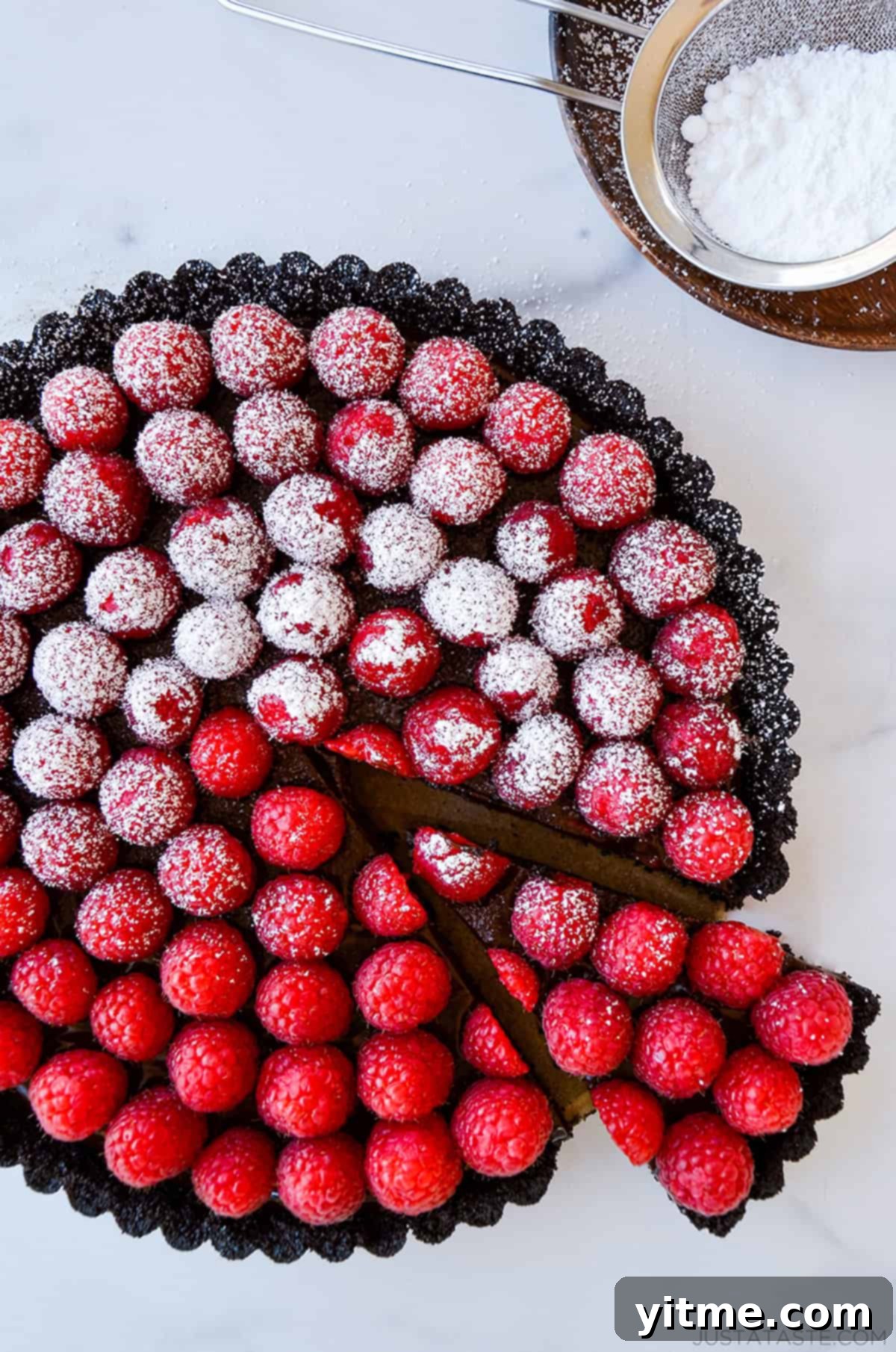 Effortless Raspberry Chocolate Tart 2 A luscious chocolate tart with a dusting of powdered sugar and topped with fresh raspberries. A slice has been removed, revealing the rich chocolate filling.