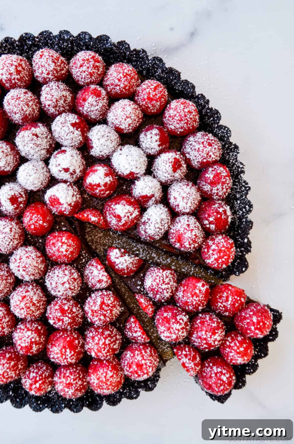 Effortless Raspberry Chocolate Tart 8 A slice is cut out of the chocolate tart, with raspberries on top and a dusting of powdered sugar.